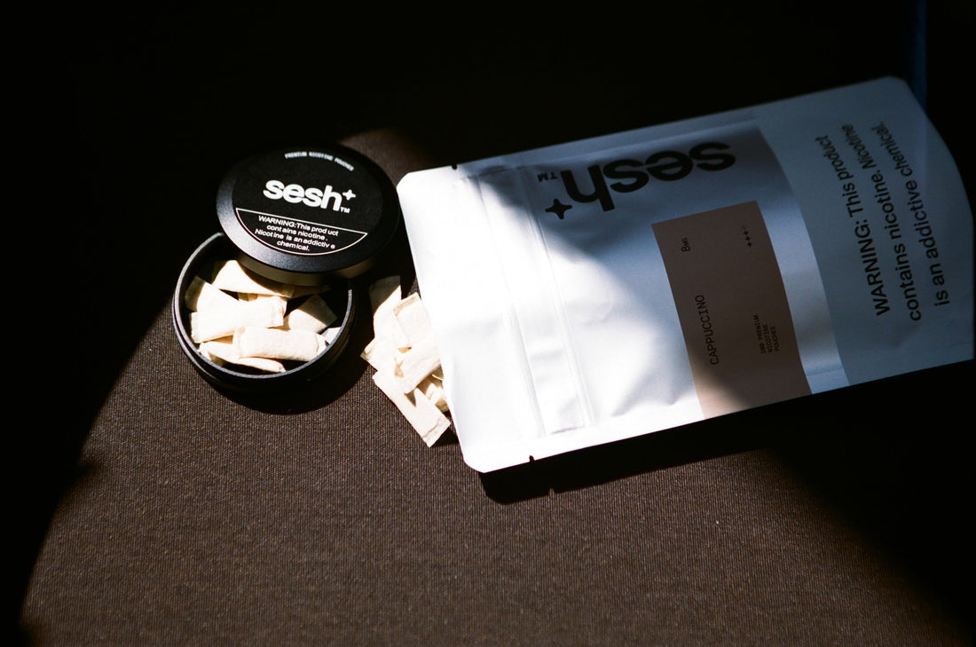 An open tin of Sesh+ Cappuccino nicotine pouches beside a foil-sealed pouch bag, lit in dramatic side light.